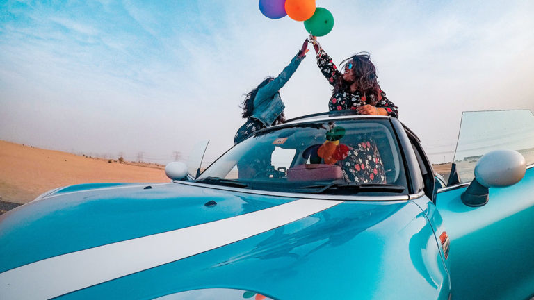 Two people joyfully reach for colorful balloons while standing in a bright blue convertible under a clear sky, conveying a sense of freedom and adventure.