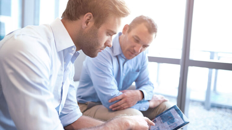 Two men in light blue shirts are focused on a tablet screen in a bright office. The mood is collaborative and professional, highlighting teamwork.