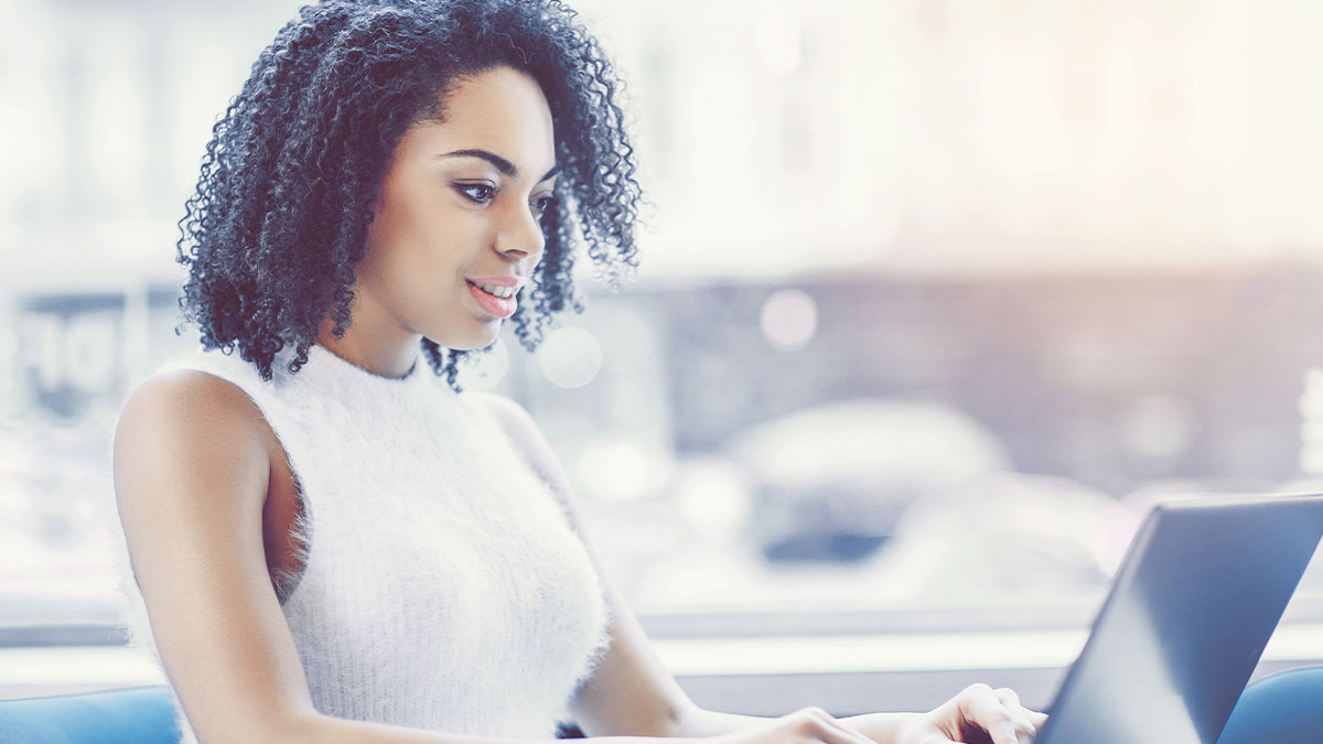 A woman with curly hair and a white sleeveless top is focused on a laptop in a bright, softly blurred indoor setting, conveying concentration and warmth.