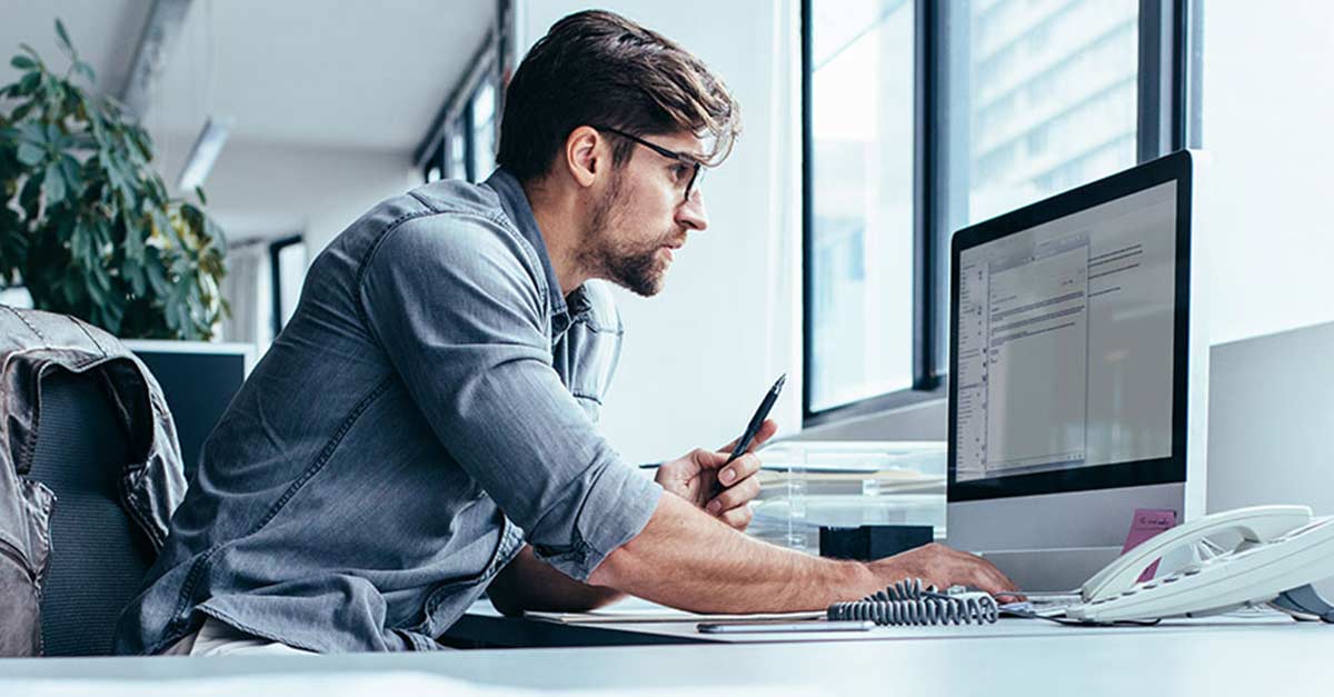 A man in a casual shirt sits at a desk in a modern office, focused on a computer screen. A jacket hangs on his chair. Natural light and plants create a calm atmosphere.