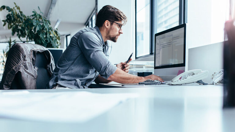 A focused man in casual attire works intently on a computer at a bright office desk. A jacket hangs on the chair; large windows provide natural light.