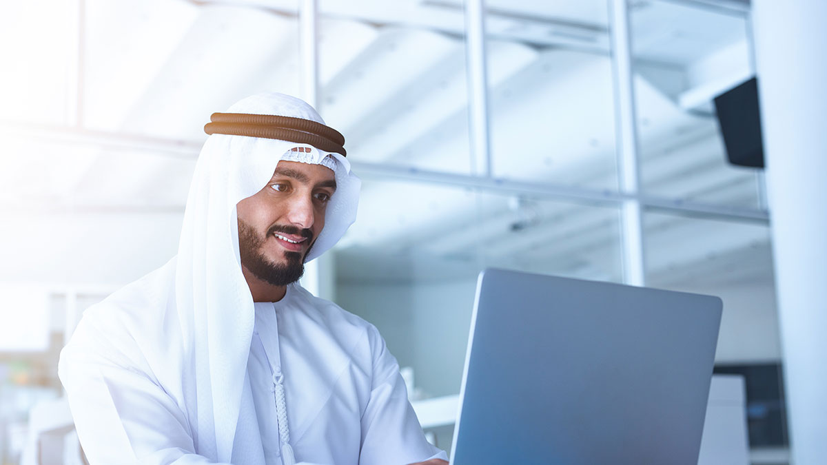 A man smiles while working on a laptop in a modern, bright office setting. The atmosphere is professional and focused.