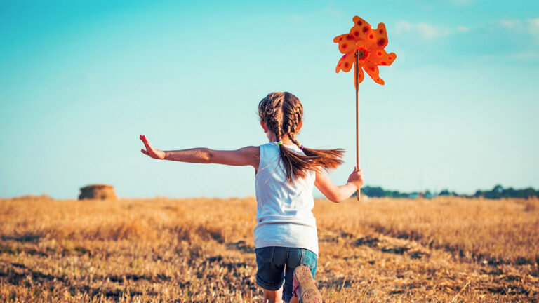 A girl with braided hair runs through a sunlit field, holding an orange pinwheel. Her arms are outstretched, capturing a sense of freedom and joy.