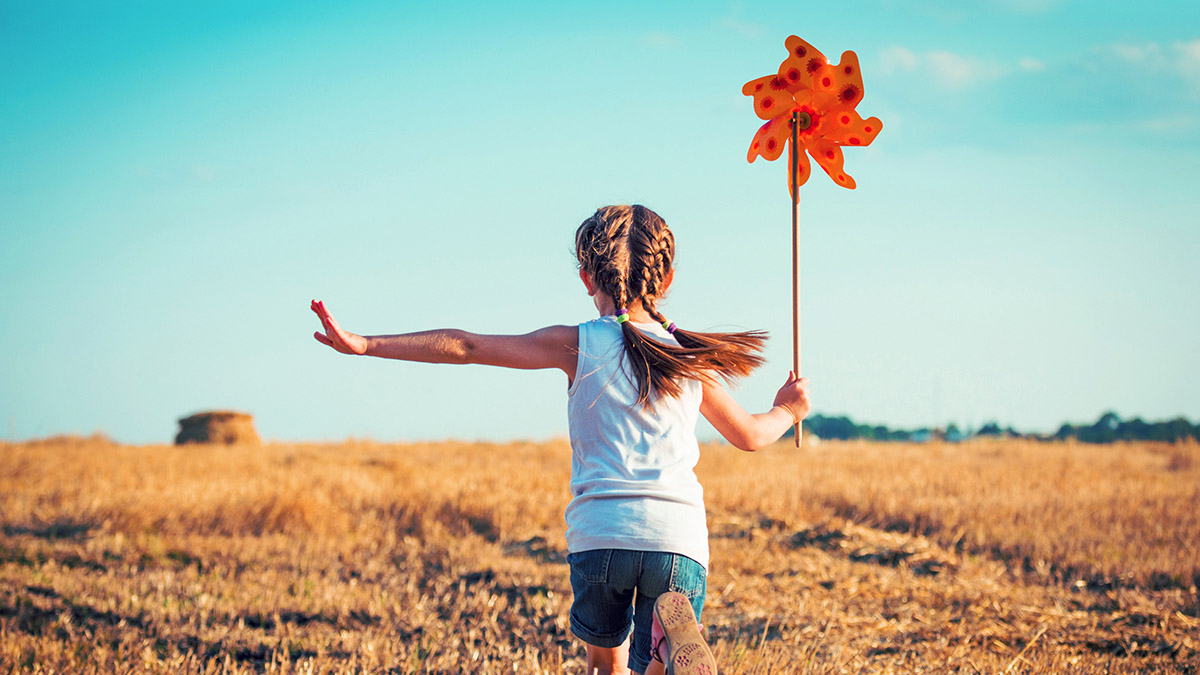 A girl with braided hair runs through a sunlit field, holding an orange pinwheel.