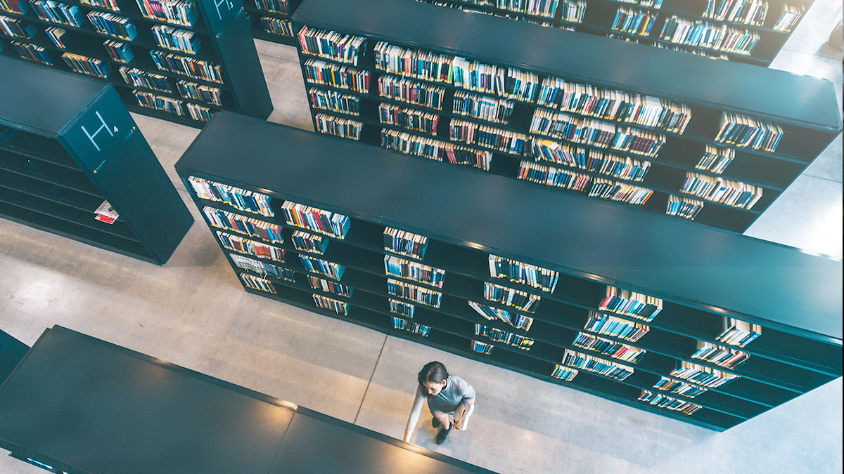 Aerial view of a library with rows of black bookshelves filled with colorful books. A person walks between aisles, creating a quiet, contemplative atmosphere.