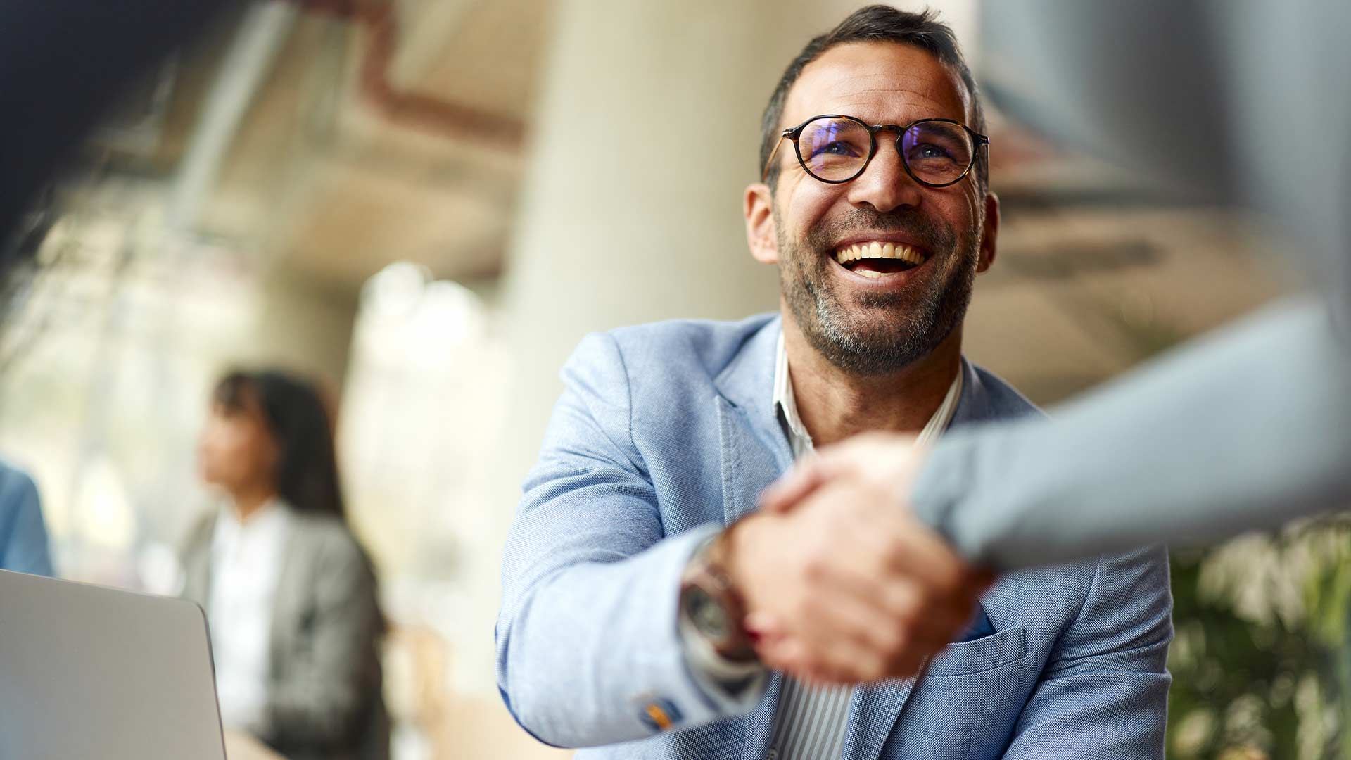 A man in glasses and a blue blazer smiles warmly while shaking hands with another person, conveying a friendly business interaction in an office setting.