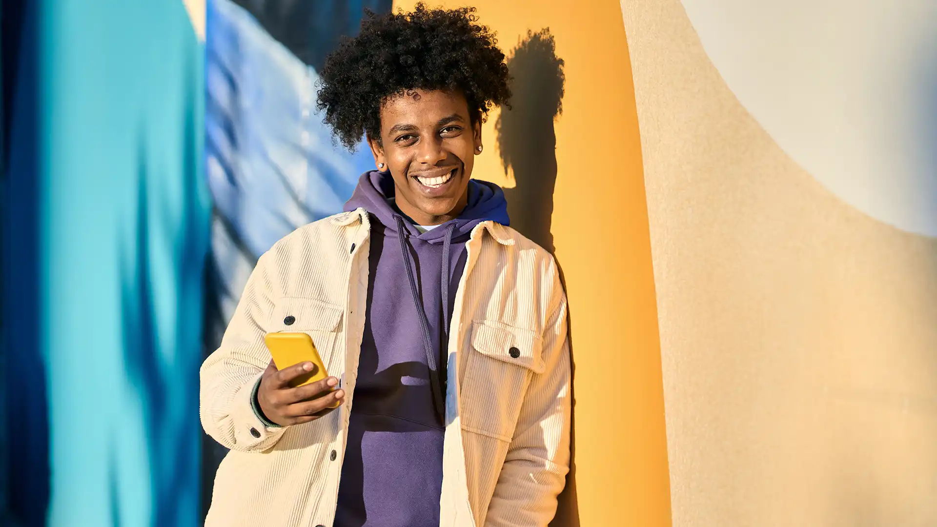 Young man leaning on wall holding yellow cell phone and smiling looking at camera