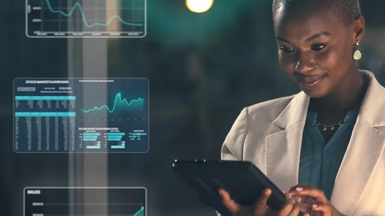 On the right a woman in dark green shirt, with khaki blazer on, holding a black tablet in her right hand while tapping screen with another. On the left side of the image there is super imposed charts and graphs, implying this is what's on the woman's screen.