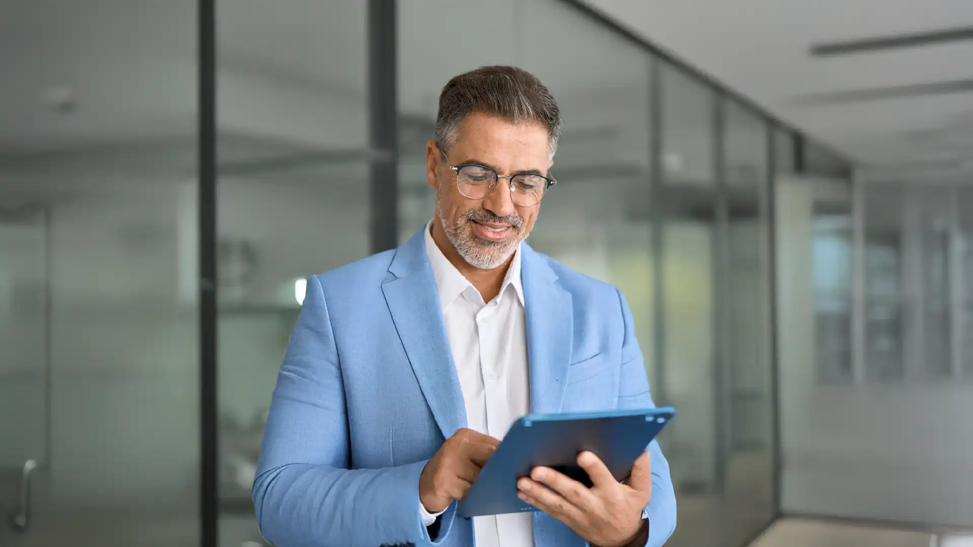 Man in blue suit jacket holding tablet in conference room that's made with glass walls. 